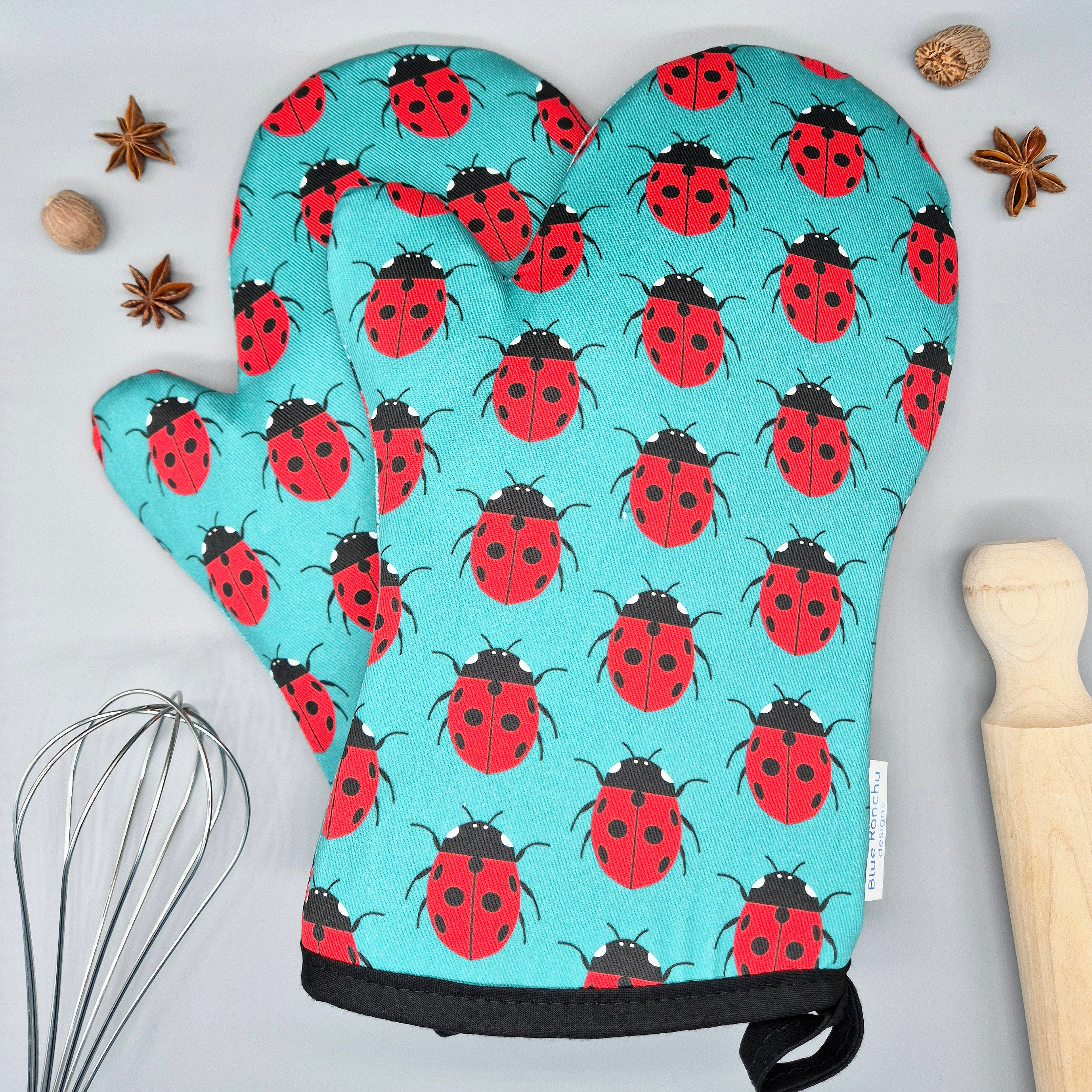 Pair of oven mitts with ladybird pattern on a turquoise background, surrounded by kitchen items.