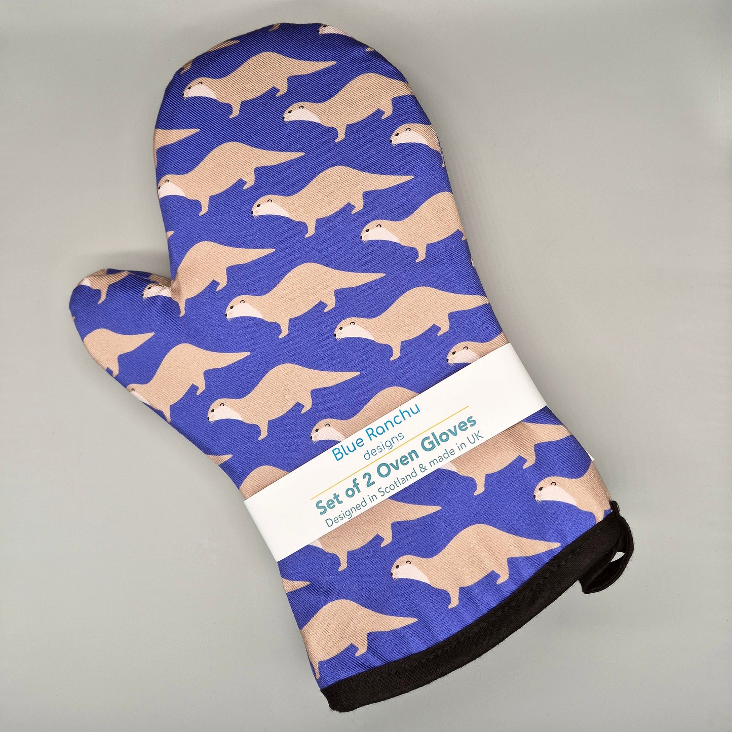 Oven mitts with otter pattern on a blue background, displayed on a gray surface.