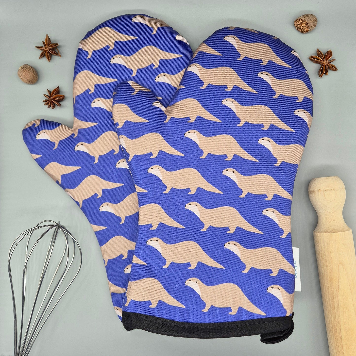 Oven mitts with otter pattern on a blue background, placed on a gray surface with kitchen items.