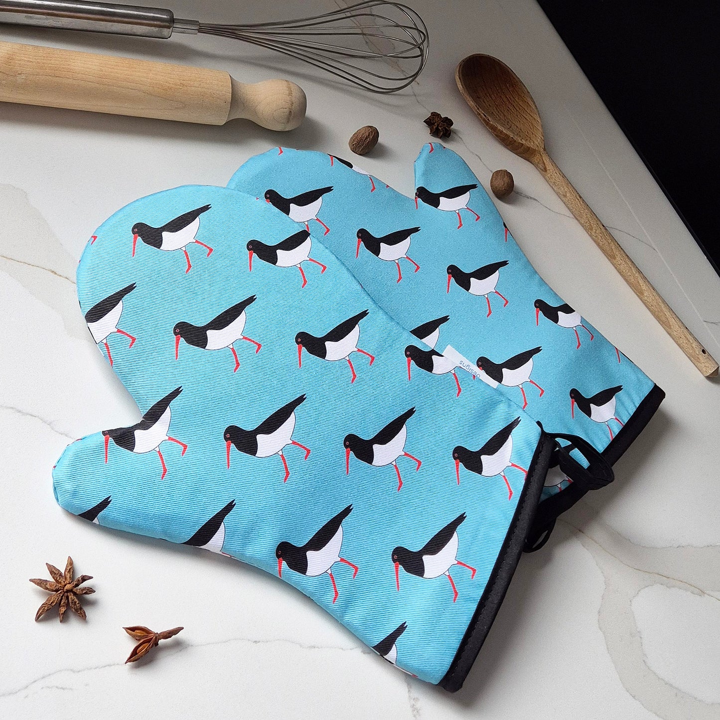 Pair of blue oven mitts with Oystercatcher bird pattern on a marble surface with kitchen tools.