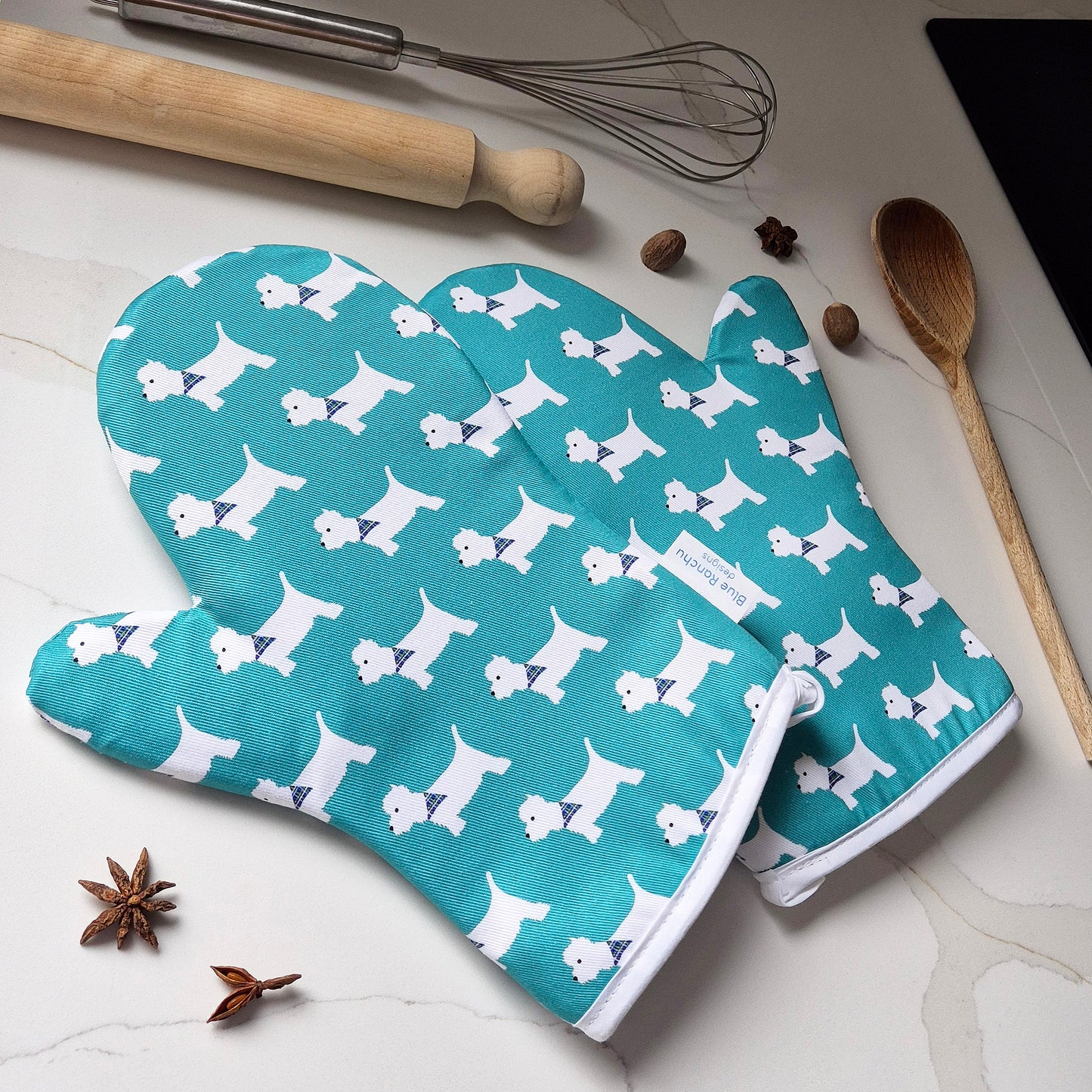 Pair of turquoise oven mitts with white Westie dog pattern on a kitchen counter.