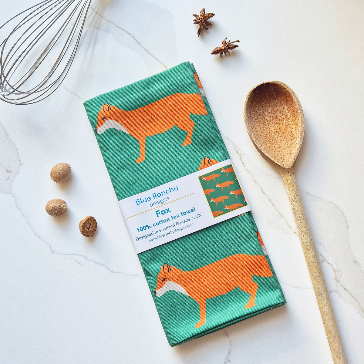 A folded cotton tea towel with a green background and orange fox pattern, wrapped in a paper band labeled 'Blue Ronchu Designs Fox'. The towel is styled in a flat-lay on a white marble surface with a wooden spoon, a wire whisk, and star anise.