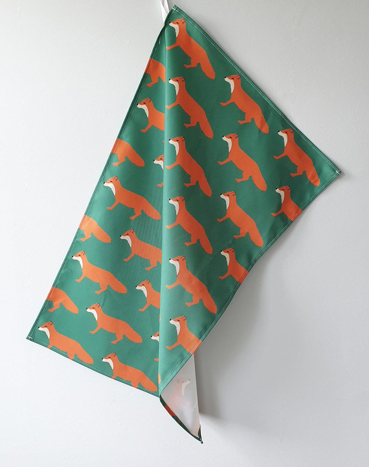 Tea Towel with a repeating pattern of orange foxes on a dark green background, hanging against a plain light gray wall.