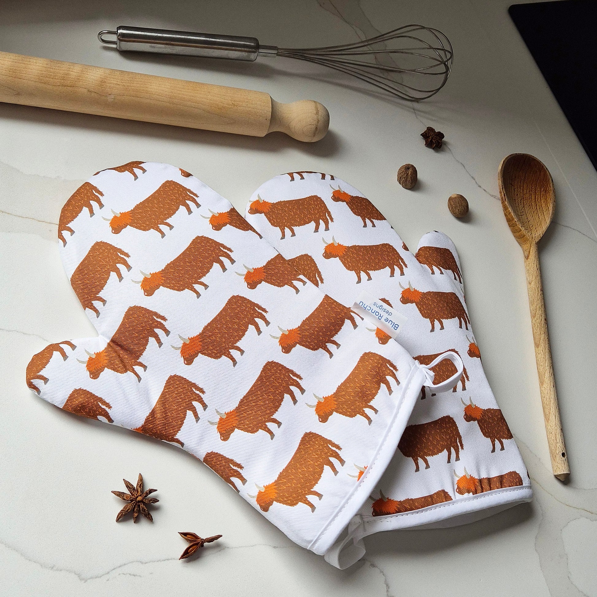 Oven mitts with Highland cow pattern on a kitchen counter with utensils and spices.