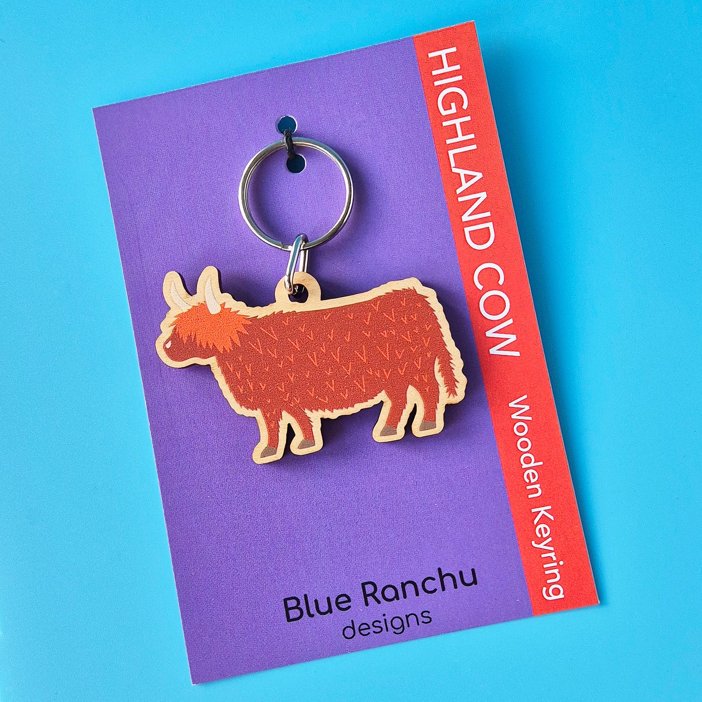 Highland cow wooden keyring on a purple card with 'Blue Ranchu designs' branding against a blue background.