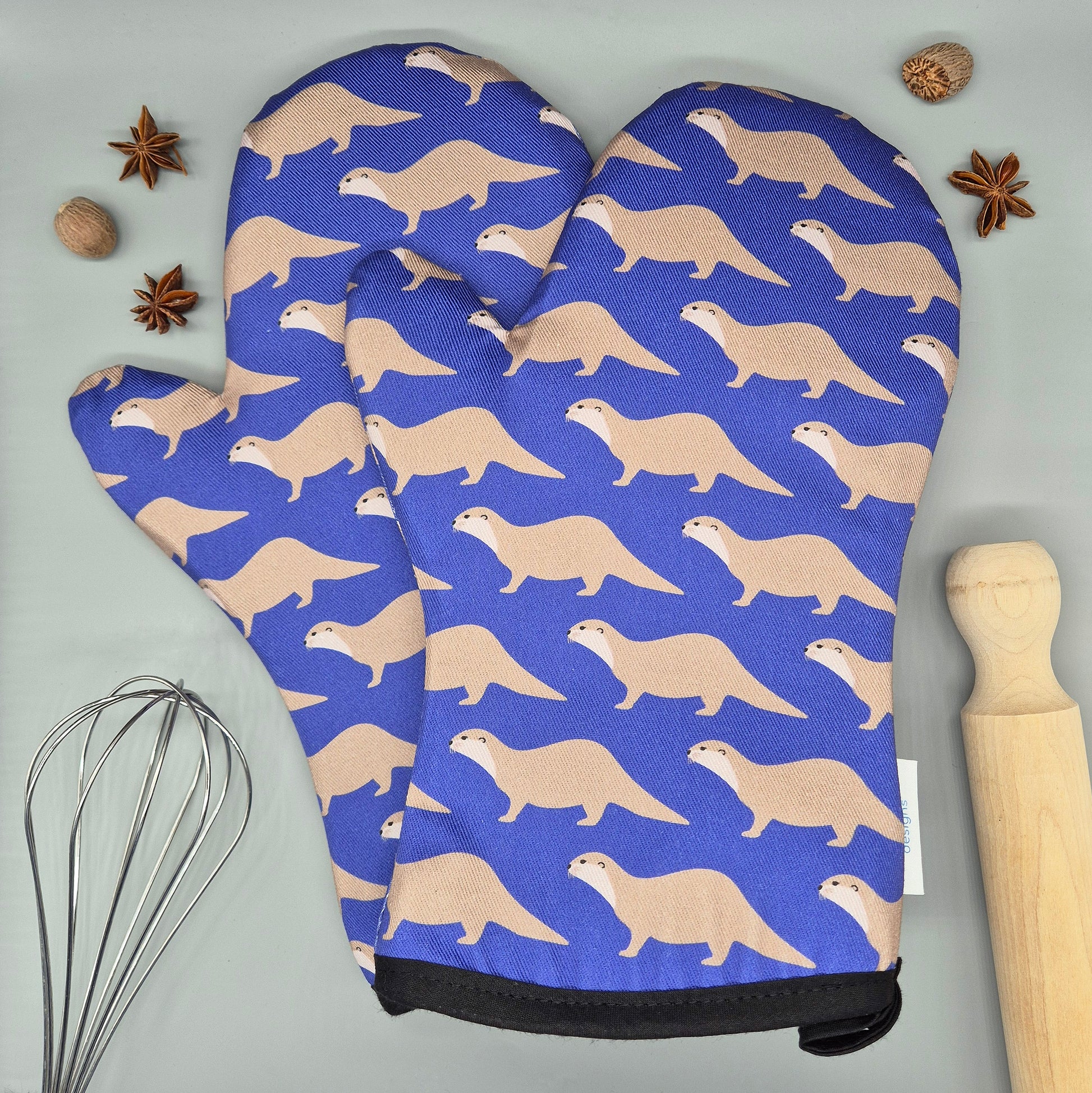 Oven mitts with otter pattern on a blue background, placed on a gray surface with kitchen items.