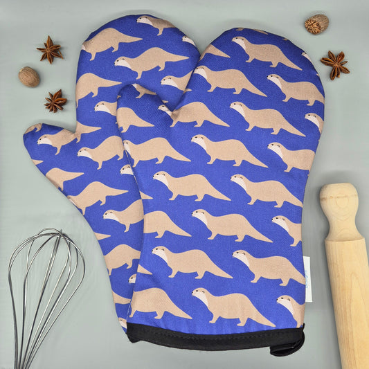 Oven mitts with otter pattern on a blue background, placed on a gray surface with kitchen items.