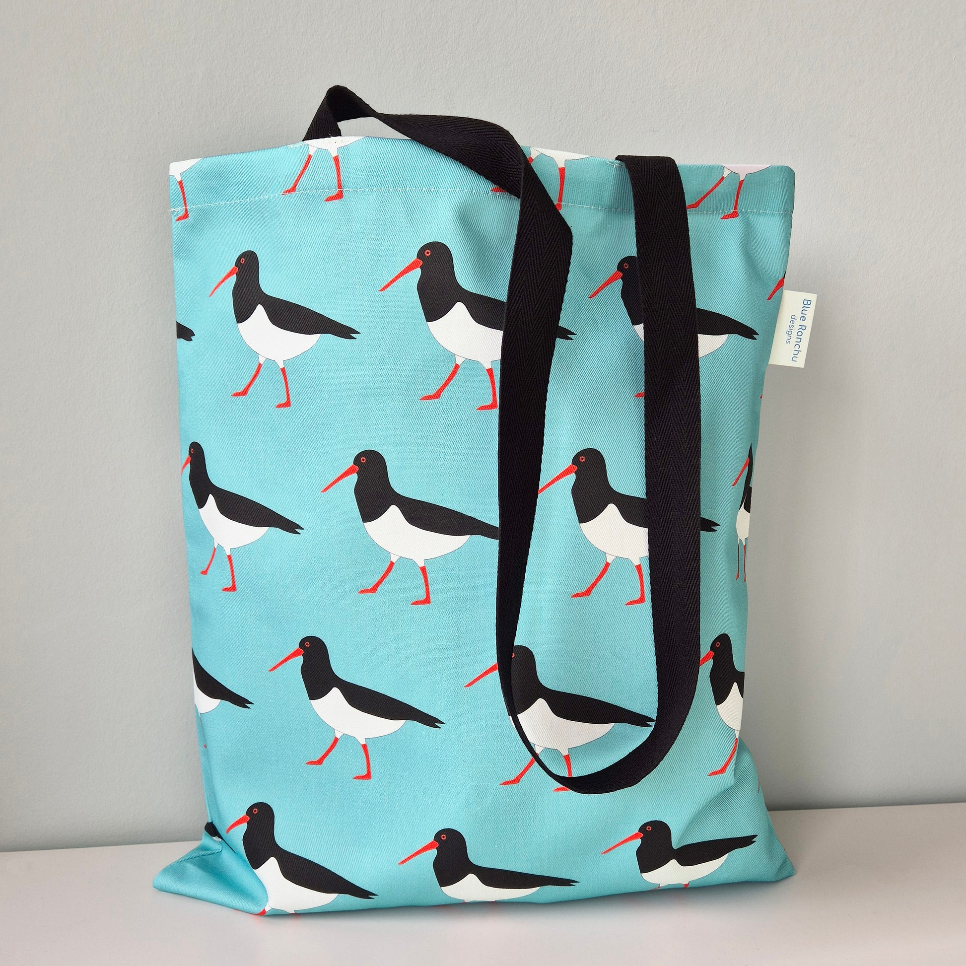 Tote bag with an Oystercatcher pattern and black handles on a turquoise background