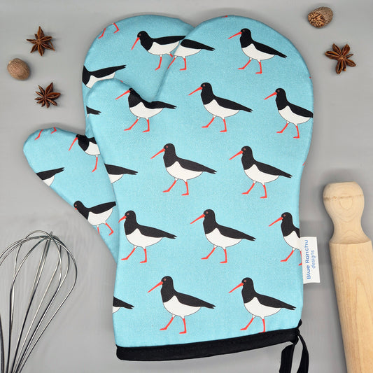 Oven mitts with Oystercatcher bird pattern on a light blue background, placed on a gray surface with kitchen items.