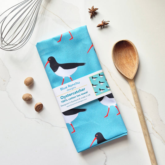 A folded cotton tea towel with an oystercatcher print is laid flat on a light marble surface, wrapped with a paper belly band. The towel features black and white birds with red-orange beaks and legs on a bright blue background.
