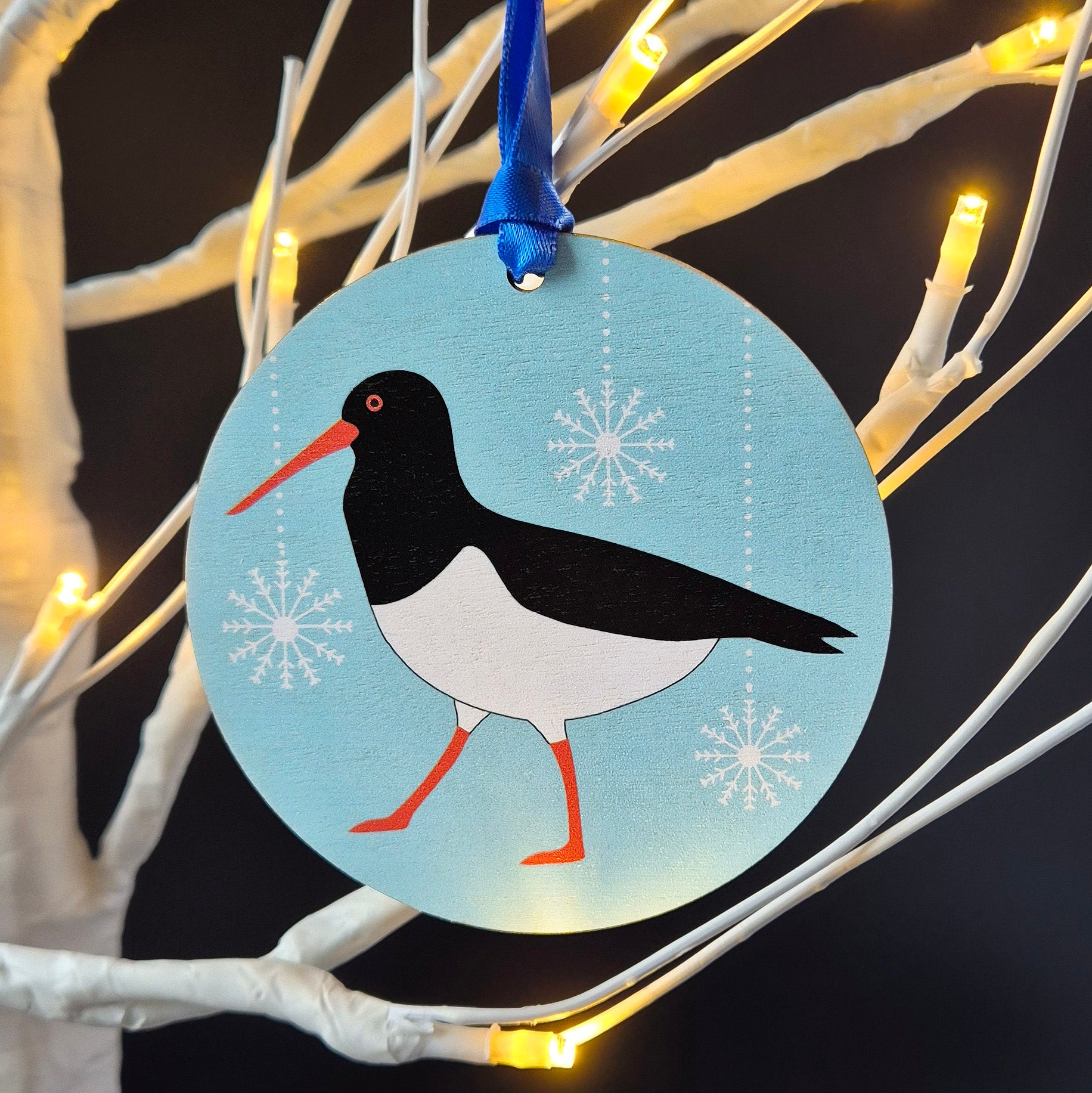 Decorative wooden ornament with an Oystercatcher design hanging on a branch with lights.