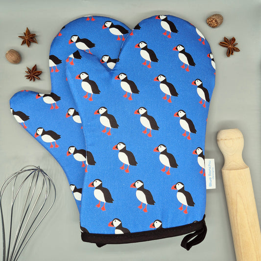 Pair of blue oven mitts with puffin pattern on a gray surface with kitchen items.