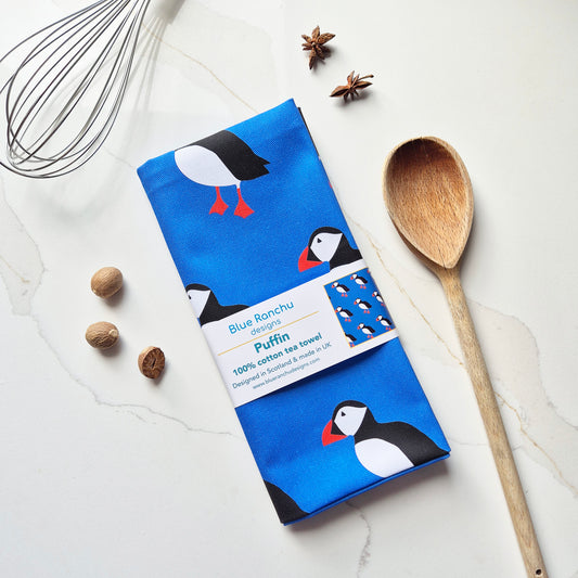 A packaged kitchen towel with a puffin print, laid flat on a light marble surface with a wooden spoon, whisk, star anise, and whole nuts as styled props. The towel label reads Blue Ronchu Designs and Puffin.