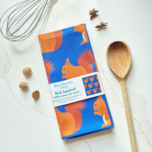 A Blue Ranchu Designs tea towel with a 'Red Squirrel' pattern, styled in a flat-lay on a white marble surface with a wooden spoon, nuts, star anise, and a wire whisk.