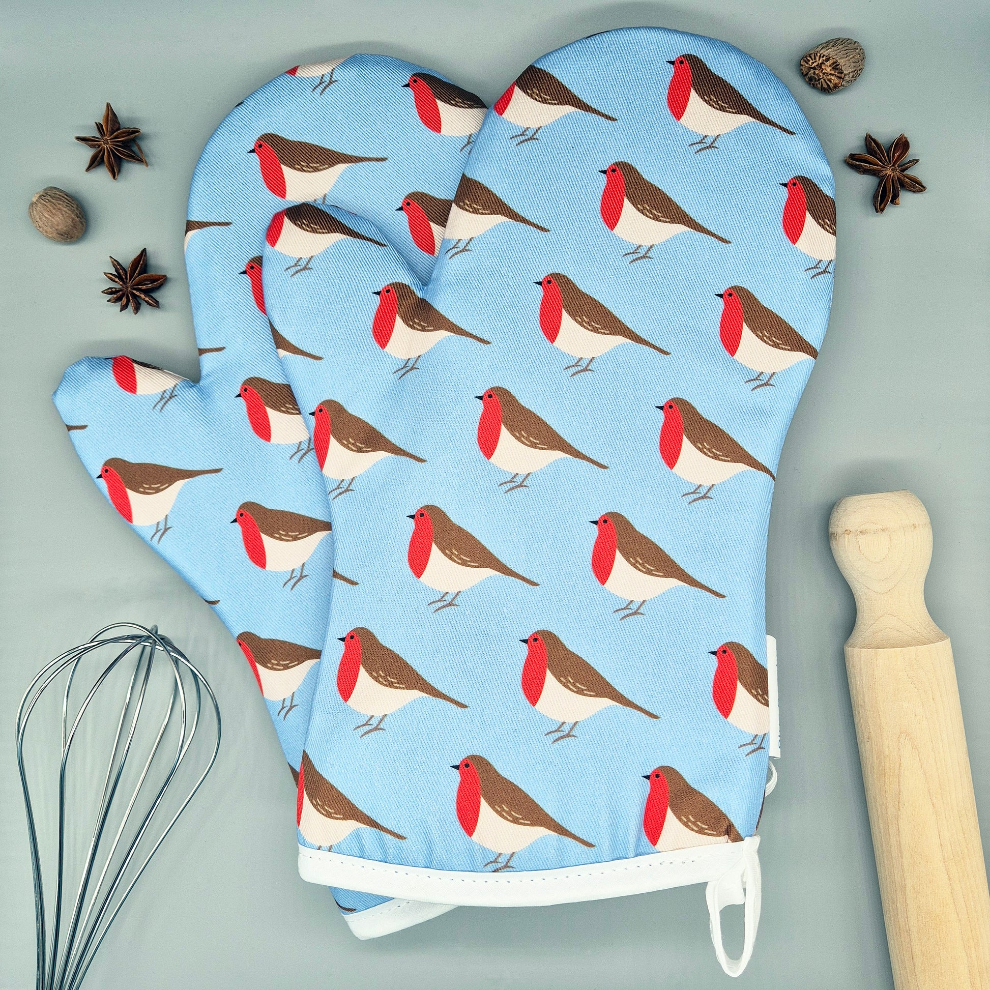 Oven mitts with robin pattern on a light blue background, surrounded by kitchen items.